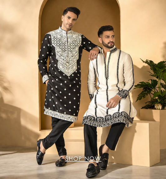Two men in traditional black and white sherwanis standing against a beige wall with a plant in the background.